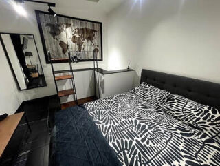 A modern bedroom with a black and white patterned bed, a large map artwork on the wall, and a standing mirror. The room has minimalistic decor and dark flooring.