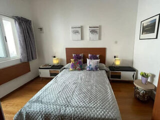 A small bedroom with a double bed, patterned bedspread, and decorative pillows. Two lamps on side tables, artwork above the headboard, a window with curtains, and a plant on the right.