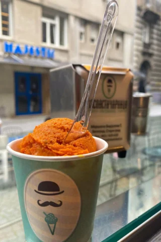 A cup of orange sorbet with a clear plastic spoon is placed on a glass surface. The cup features a logo with a mustache and bowler hat. A building is in the background.