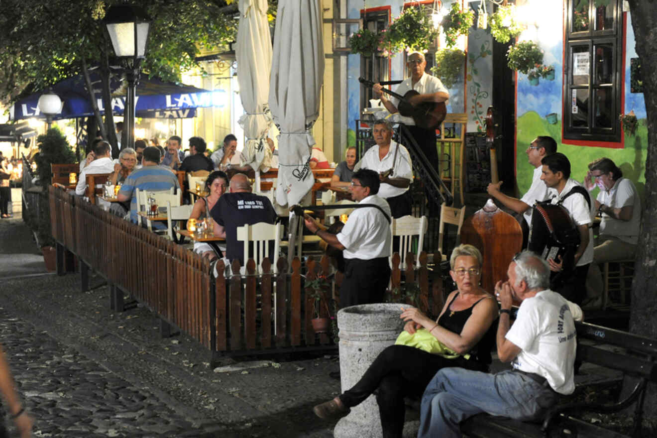 Outdoor restaurant scene with people dining and musicians performing. The setting has a rustic feel with cobblestone streets and dim lighting, creating a lively and social atmosphere.