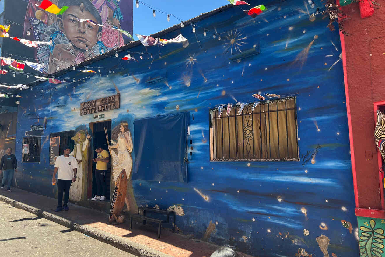 A vibrant blue mural covers a building wall with celestial designs and human figures. Colorful flags hang above, and a few pedestrians walk nearby.