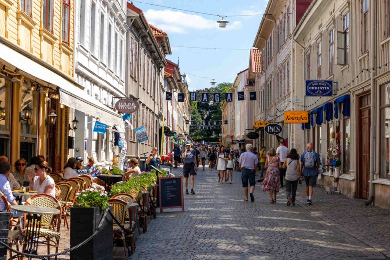 A cobblestone street with people walking and dining at outdoor cafes. Shops with colorful signs line both sides.