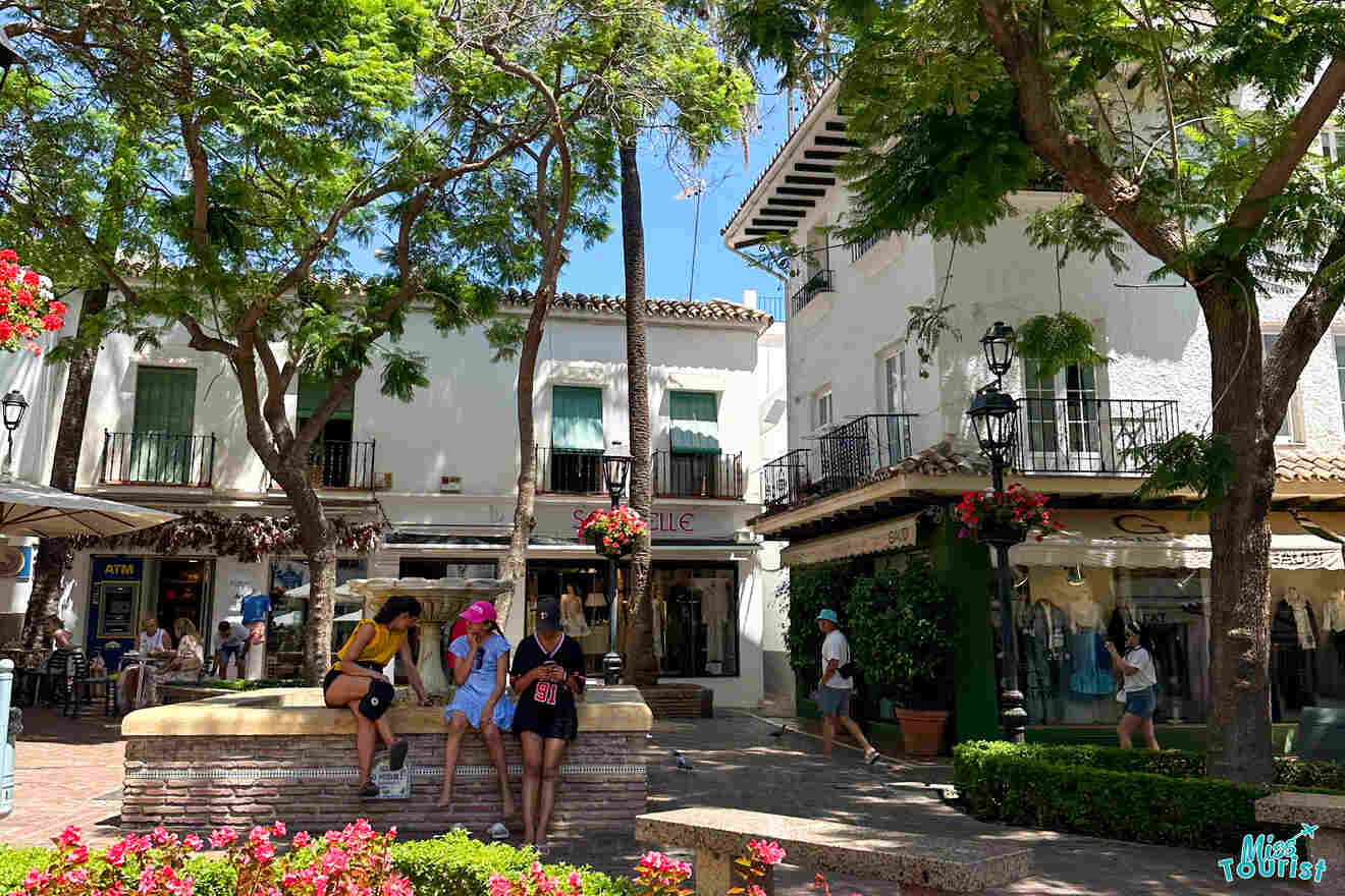 People sitting on a raised platform surrounded by trees in a sunny plaza with nearby shops.