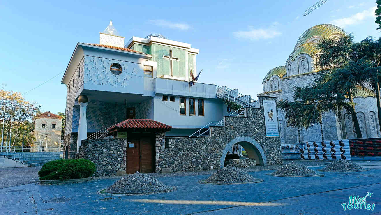 Stone building with religious symbols and a cross, arched entry, steps leading up, and piles of stone in front. Another stone building with a dome is visible in the background.