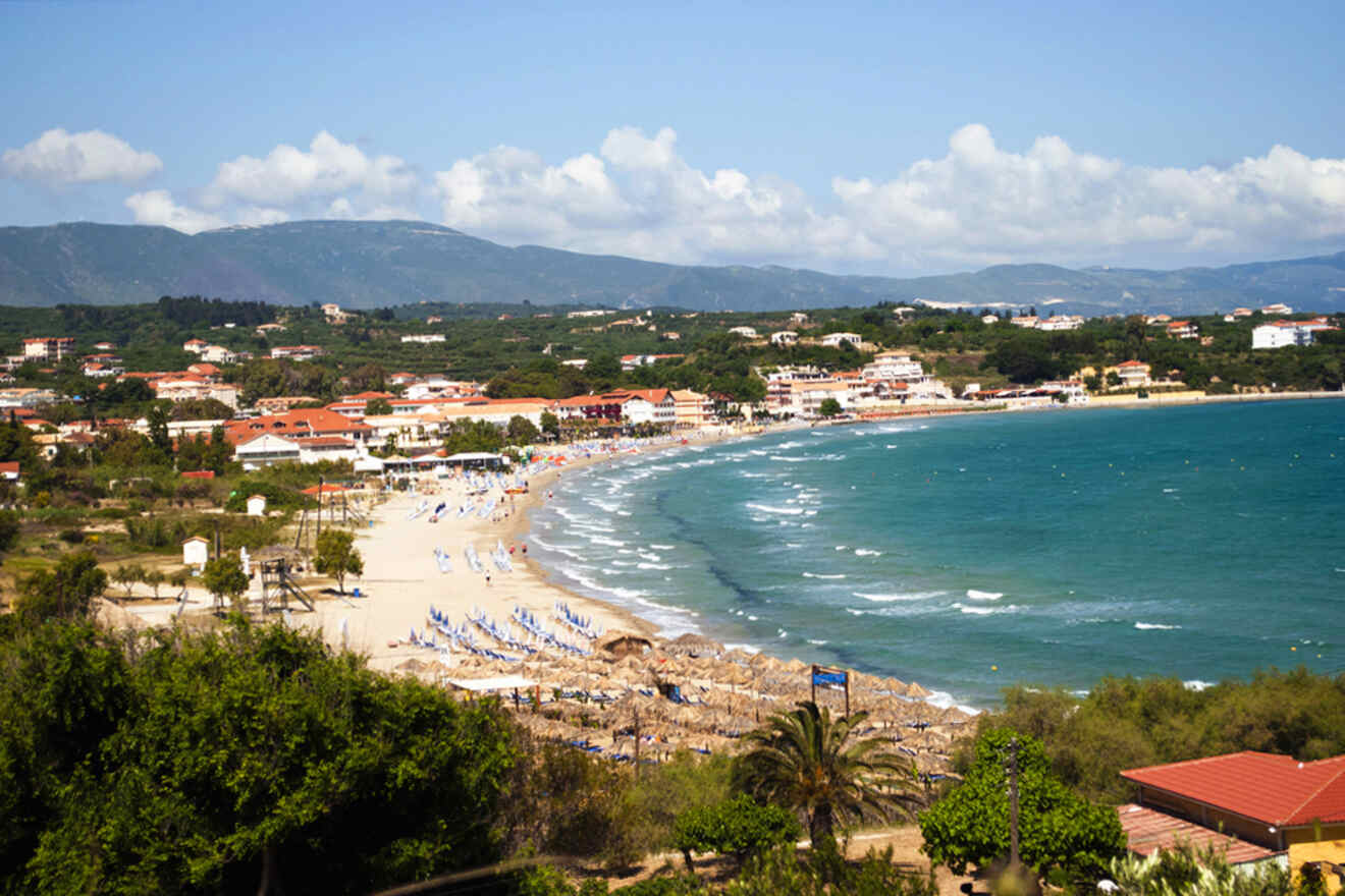 A scenic view of a coastal town with a sandy beach, clear water, umbrellas, and lounge chairs. Hills and scattered buildings are visible in the background under a partly cloudy sky.