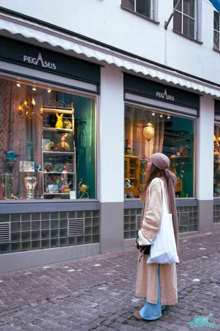 author of the post in a long coat and headscarf stands in front of a store window display on a cobblestone street.