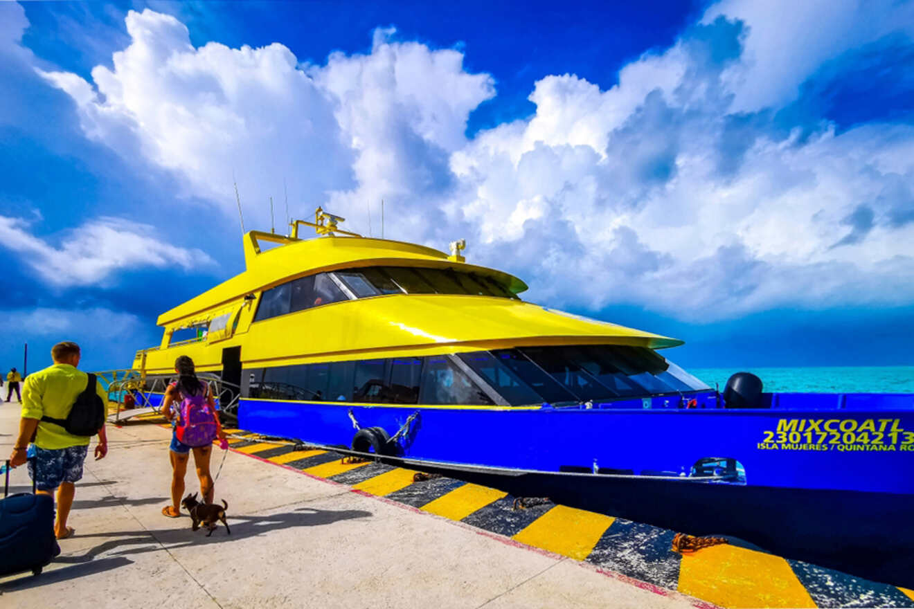 A large yellow and blue ferry docked at a pier with two people, a dog, and rolling luggage walking towards it under a partly cloudy sky.