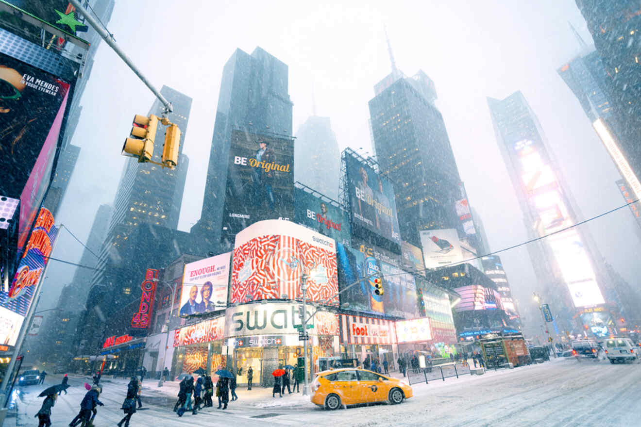 Times Square in heavy snowfall; bright billboards illuminate the scene, coated buildings, and streets. People walk while a yellow taxi drives past, and traffic lights are visible.