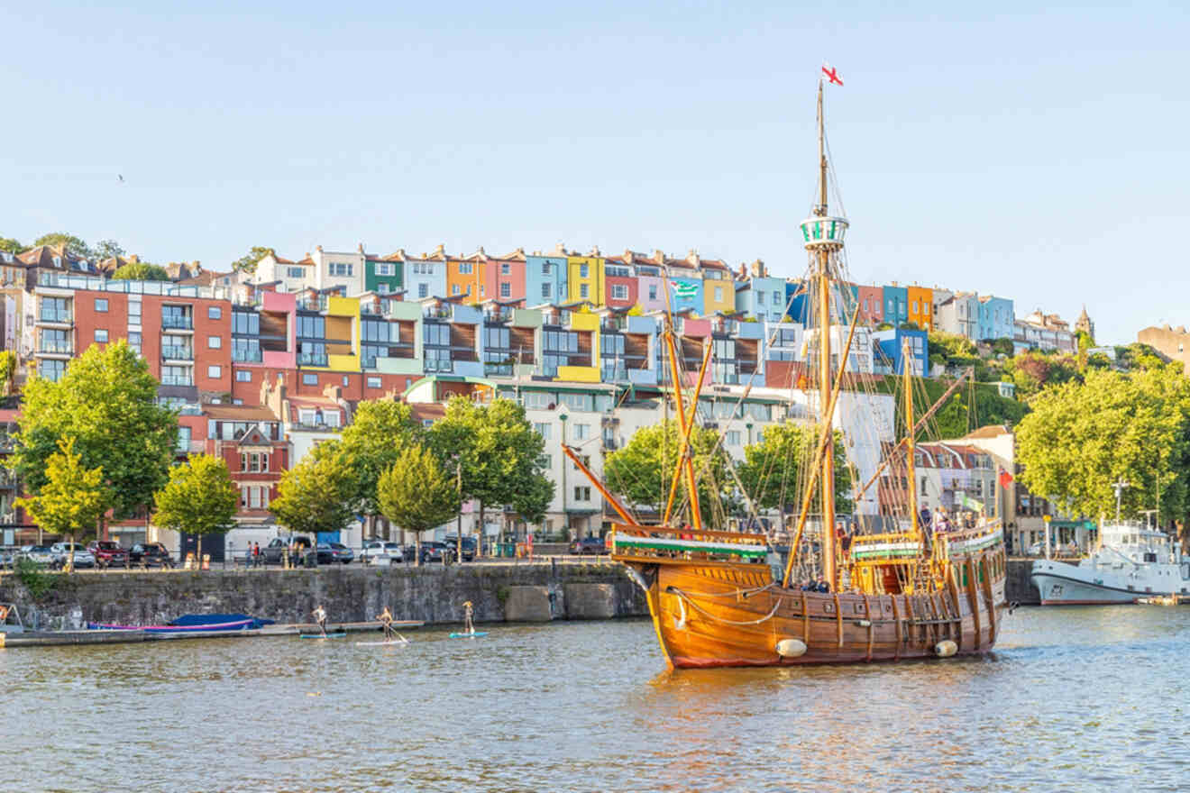 A wooden ship sails in a harbor with colorful houses on a hillside in the background. Trees line the waterfront.