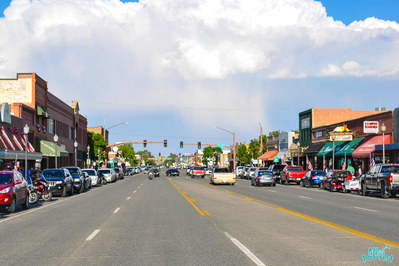 A busy small-town street with parked cars and storefronts on both sides under a cloudy sky. Traffic lights are visible in the distance.