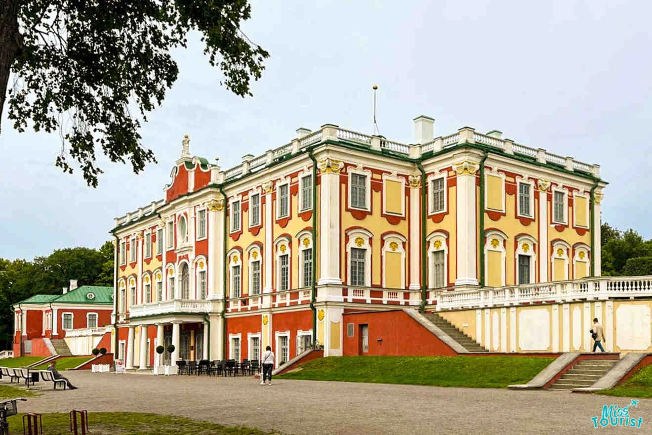 A large, ornate red and yellow baroque-style building with white trim. People walk nearby. The sky is overcast, and there's a tree on the left.