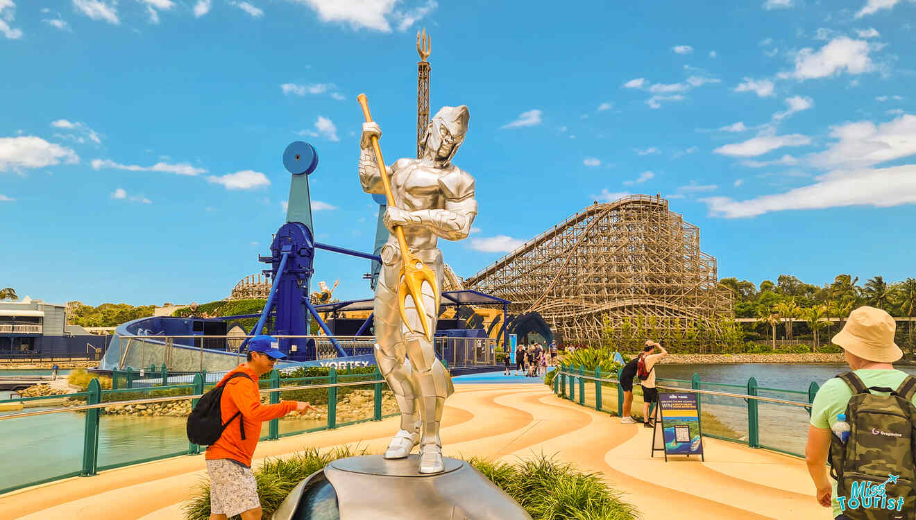 People walking near a silvery statue of a superhero, with a wooden roller coaster and a blue ride in the background under a clear sky.