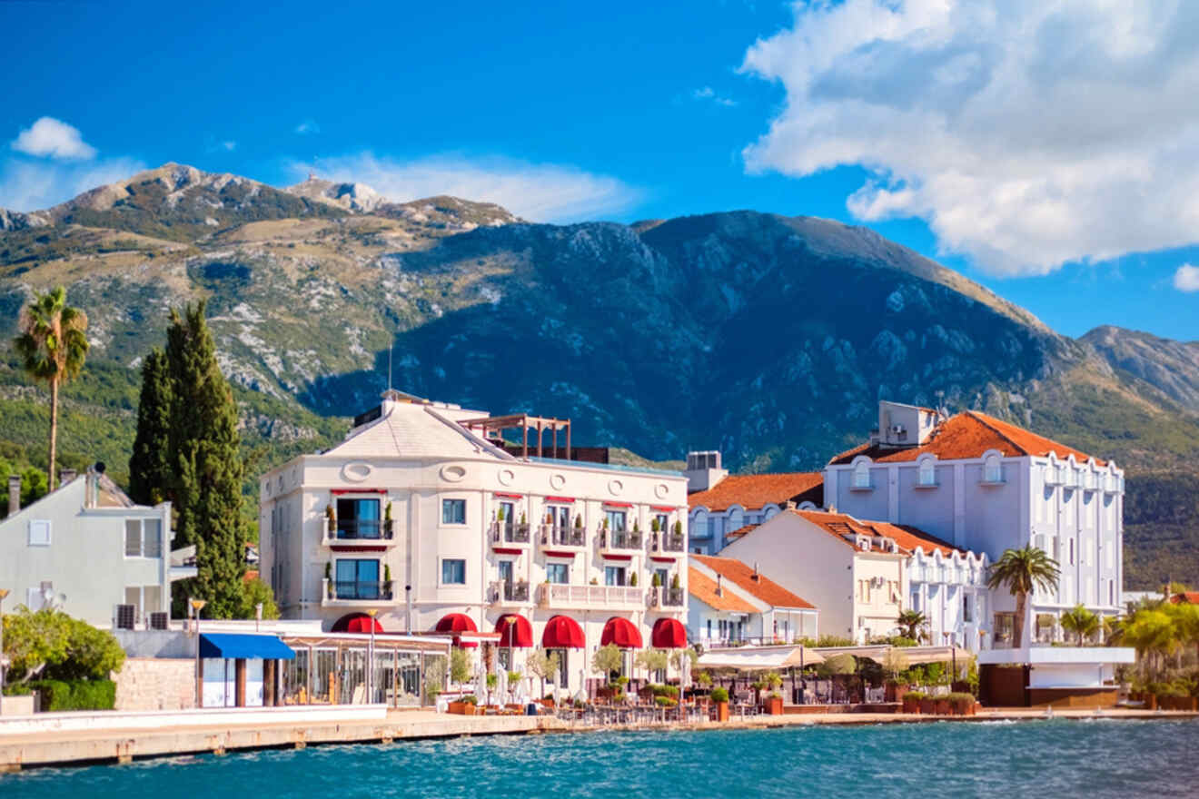 Scenic view of coastal buildings with red roofs by the water, framed by mountains and a clear blue sky.