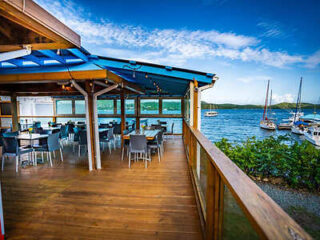 Outdoor restaurant deck overlooking a lake with sailboats, featuring wooden flooring, blue roof, and dining tables with chairs under a clear blue sky.