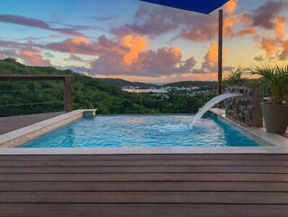 Infinity pool with a water feature overlooks lush green hills and a distant body of water under a colorful sunset sky.