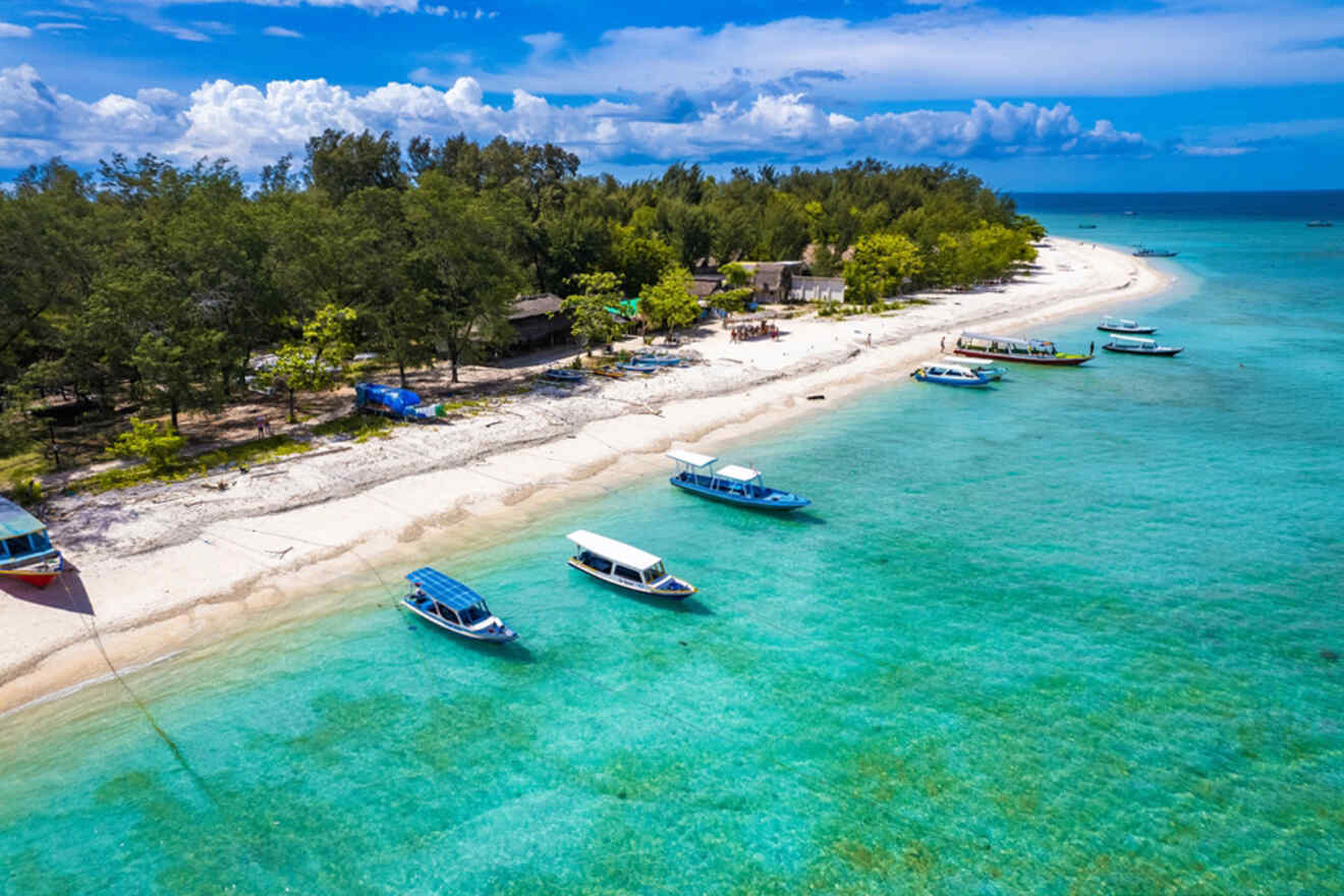 Aerial view of a sandy beach with anchored boats on clear turquoise water, surrounded by lush green trees and a blue sky.