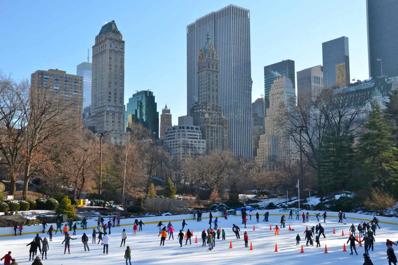 People ice skating on a rink surrounded by tall buildings and trees in a cityscape under a clear blue sky.