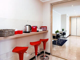 Modern kitchen area with red bar stools, a wooden counter holding a kettle, coffee machine, and basket; small dining tables visible in the adjoining room.