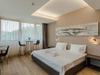 Hotel room with a neatly made double bed, two bar stools by a window, a desk, and wall art above the bed. Soft lighting and wood flooring create a cozy atmosphere.