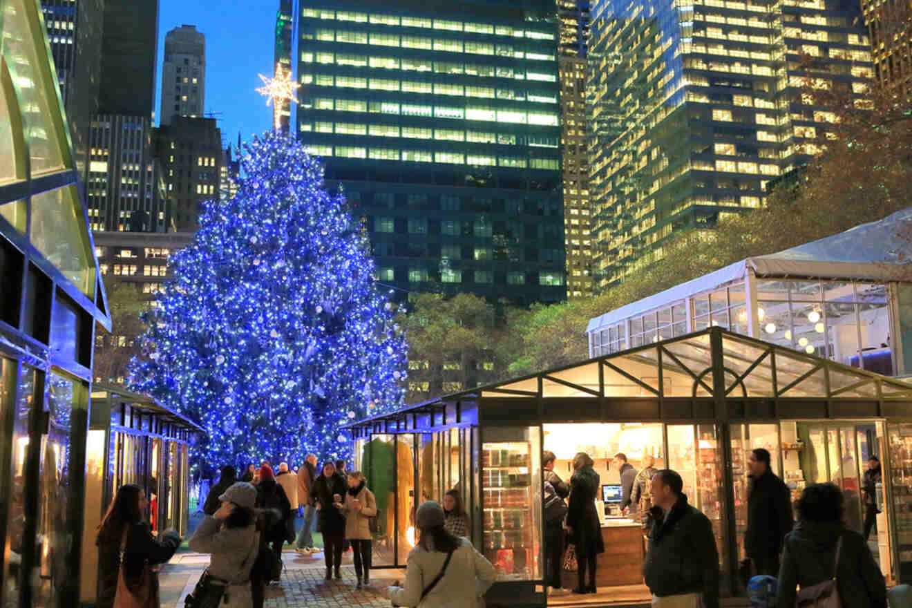 People walk through a holiday market with a large, illuminated Christmas tree in the background, surrounded by tall buildings at dusk.