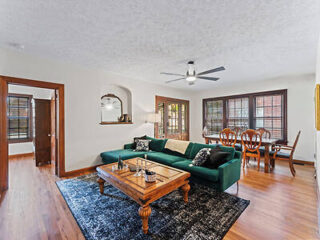 Living room with a green couch, wooden coffee table, ceiling fan, and dining area. Hardwood floors and large windows provide natural light.