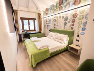 Cozy bedroom with a green bed and colorful bubble-patterned wall. Features a window, desk, and light wood flooring.