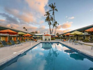 A hotel pool with lounge chairs and umbrellas on either side, surrounded by two-story buildings and tall palm trees under a partly cloudy sky.