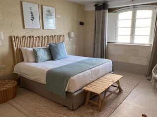 A neatly made bed with blue accents, a wooden bench at the foot, two framed leaf prints above, and a window with open shutters in a neutral-toned bedroom.