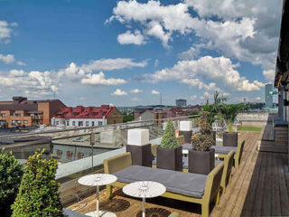 Rooftop patio with seating and potted plants, offering a view of buildings and a cloudy sky.