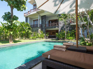 Outdoor pool with lounge chairs and umbrella in front of a two-story house surrounded by lush greenery.