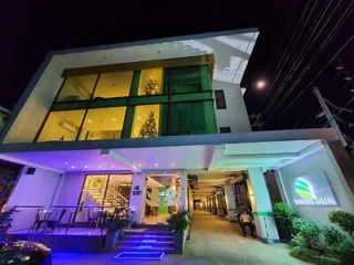 Night view of a modern hotel with illuminated windows and a visible Christmas tree inside. The entrance is well-lit, featuring a logo sign and a small outdoor seating area.