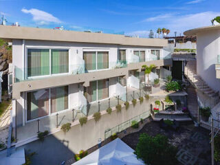 Modern two-story building with large glass windows, a balcony, and a landscaped courtyard. Clear blue sky and some greenery are visible.