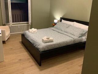 A neatly made double bed with folded towels on top, beside a small bedside table with a lamp, in a room with light green walls and wooden flooring.