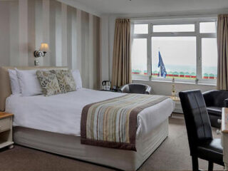 Hotel room with a double bed, striped headboard, two armchairs, a small table, and a window with a sea view. A framed flag is outside the window.