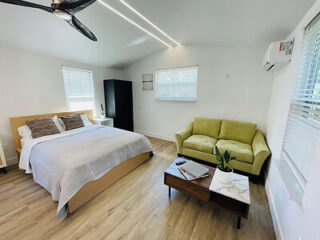 A bedroom with a bed, green sofa, wooden coffee table, black wardrobe, and white walls. Ceiling fan, air conditioner, and natural light from windows are visible.