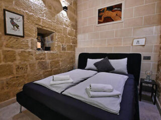 A cozy bedroom with a stone wall features a neatly made bed, two folded towels, and framed art. A small window is visible to the left.