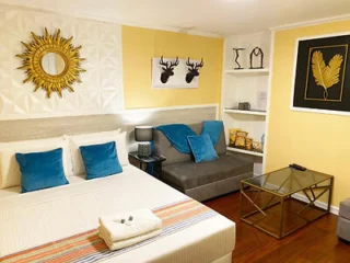 A neatly arranged bedroom with a bed, blue cushions, towels, a sunburst mirror, wall art, and a gray sofa. Shelves hold decor items, and a glass coffee table is in front of the sofa.