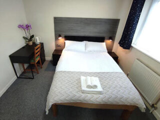 A tidy bedroom with a double bed, two bedside tables, folded towels on the bed, a small desk, a chair, a kettle, and a potted orchid flower on the desk. A window with curtains is on the right.