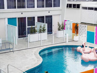 A modern outdoor pool area with inflatable pink flamingo floats. The pool is surrounded by a white tiled patio and colorful wall panels near a building with large windows.