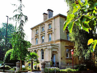 A large, beige three-story house with a classical design, surrounded by trees and a garden, under a clear sky.