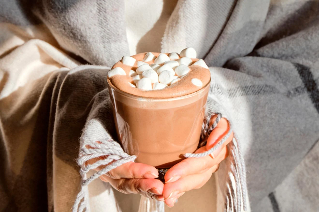 Person wrapped in a cozy blanket holding a glass of hot chocolate topped with marshmallows.