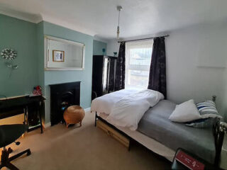 Bedroom with a bed, desk, chair, fireplace, and window. Walls are light blue and white with dark curtains. A round wicker basket is near the fireplace.