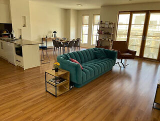 Spacious living area with a teal sofa, small side table with a plant, dining table with six chairs, brown lounge chair, and large windows. Kitchen counter with stools in the background.