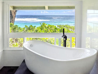 Freestanding white bathtub with a black faucet, positioned by a large window showing a view of the ocean and greenery outside.