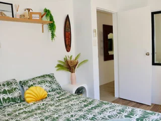 A bedroom with a green tropical-patterned bedspread, a yellow seashell pillow, wall-mounted shelf, decorative plant, and an open door leading to another room.