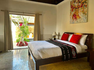 A cozy bedroom features a double bed with red and white pillows, a vibrant wall painting, and an open doorway leading to a garden with lush plants.