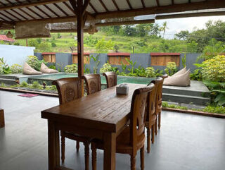 A wooden dining table with chairs is set in an open-air area overlooking a pool surrounded by greenery and bean bag seating.