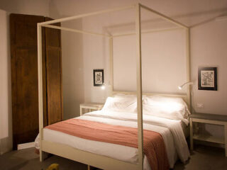 A neatly made bed with a white canopy frame, white bedding, and a pink throw, flanked by two nightstands with wall-mounted lamps and framed art above them.