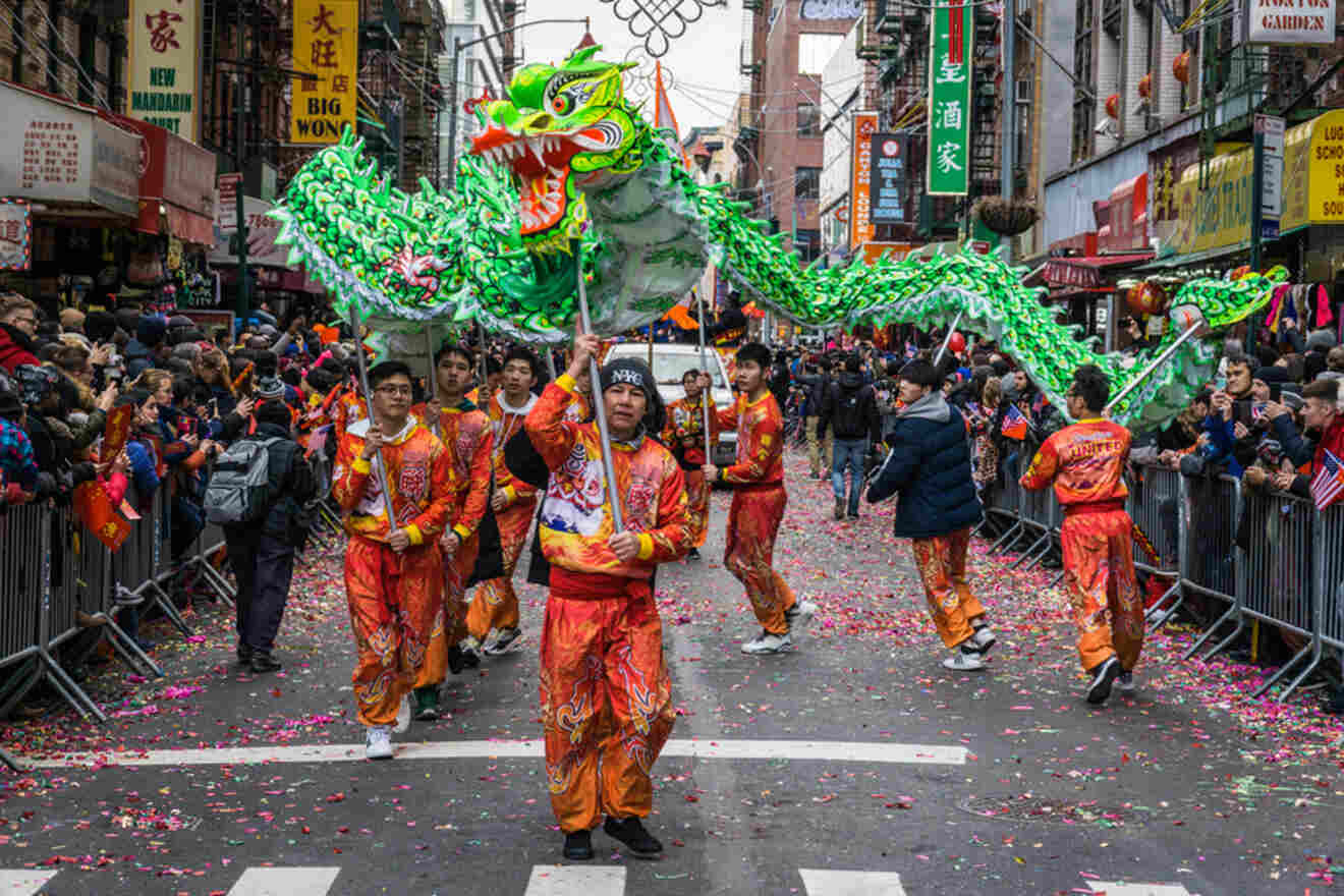 Performers in colorful costumes carry a green dragon puppet during a parade on a city street, surrounded by crowds and confetti.