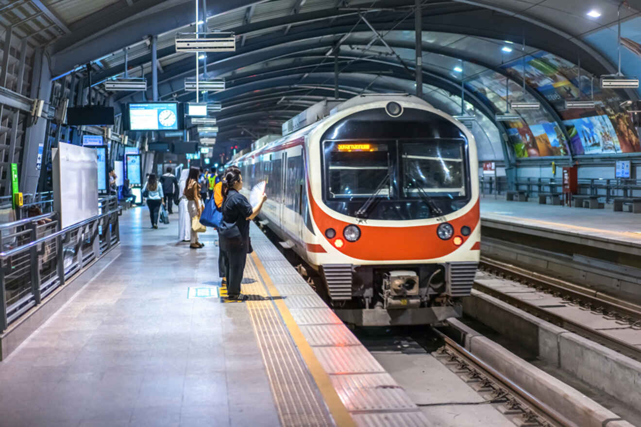 A train is stopped at a modern station, with people standing on the platform. Some are reading and others are waiting. The station is well-lit, and electronic displays are visible above.